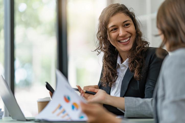 A smiling woman in business attire engaging in a friendly discussion with a colleague or client over documents and a laptop in a well-lit office setting.