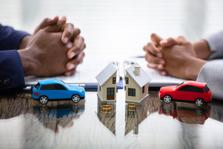 Two people with interlocked hands at a table, with toy cars and a small house model between them, symbolizing negotiations or discussions about property and automotive assets.