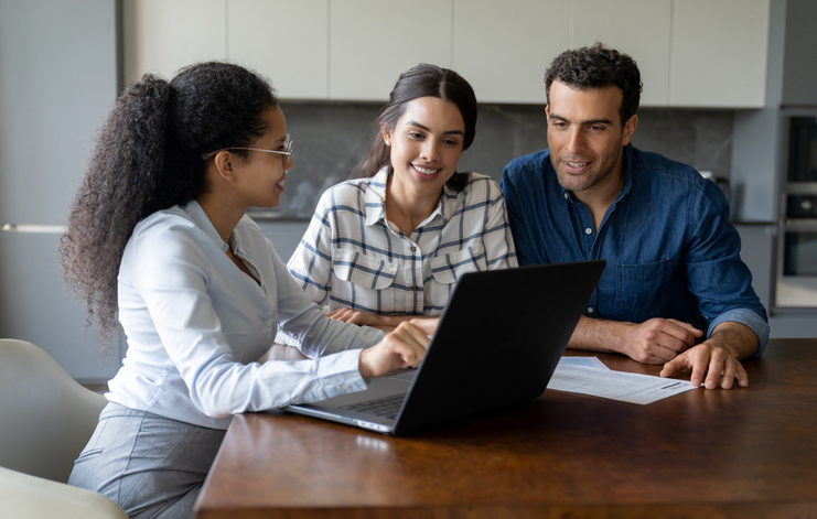 A professional woman is showing something on a laptop to a couple, who are looking on with interest, suggesting a consultation or meeting at a home or office.