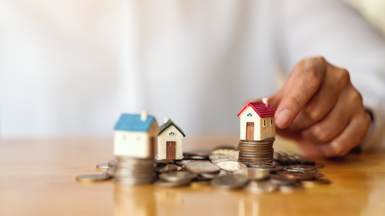 A hand is shown placing small model houses on top of stacked coins.