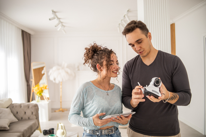 A man and woman stand close together in a bright living room, with the man holding a modern-looking security camera and the woman holding a tablet, both looking at the device with interest.